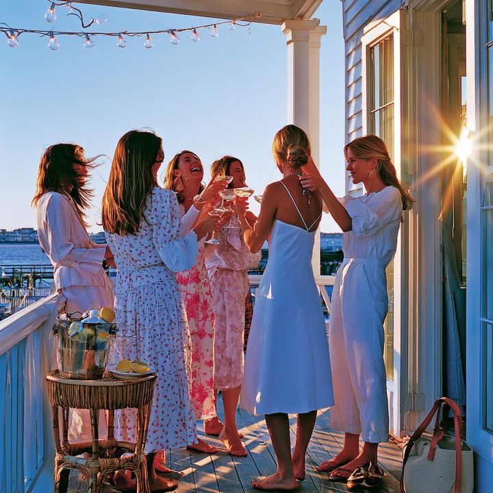 A bachelorette party in white dresses toasting at golden hour on a Charleston balcony