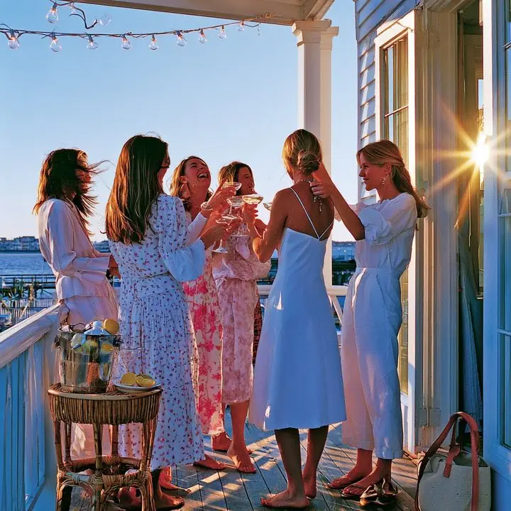 A bachelorette party in white dresses toasting at golden hour on a Charleston balcony